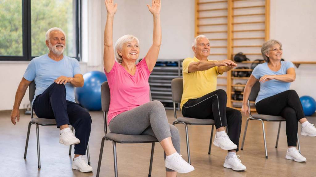 Vier ältere Menschen sitzen auf stabilen Stühlen in einem hellen Trainingsraum und machen gemeinsam Hockergymnastik. Sie führen einfache Übungen wie Armheben, Drehen des Oberkörpers und Anheben der Beine aus. Alle tragen bequeme Sportkleidung, wirken konzentriert und haben Freude an der Bewegung. Im Hintergrund sind Trainingsgeräte wie Gymnastikbälle und eine Sprossenwand zu sehen.