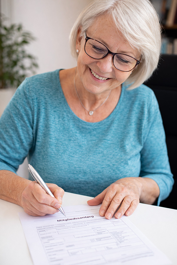 Eine ältere Frau sitzt an einem Tisch und füllt lächelnd einen Mitgliedsantrag aus. Sie hält einen Stift in der Hand und wirkt konzentriert und zufrieden. Die Umgebung ist hell und freundlich gestaltet.
